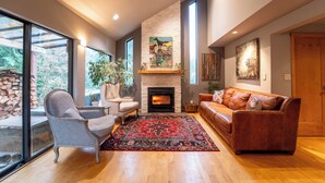 A living room with a fireplace, a sofa, a chair, and a large window. - Beautiful Farmhouse & thermal Spa in Forest (Sechelt)