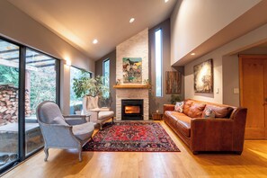 A living room with a fireplace, a sofa, a chair, and a large window. - Beautiful Farmhouse & thermal Spa in Forest (Sechelt)