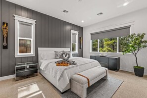 Ein modernes Schlafzimmer mit einem großen Bett, Nachttischen, einer Kommode, einem Fenster mit Blick und einer Topfpflanze.