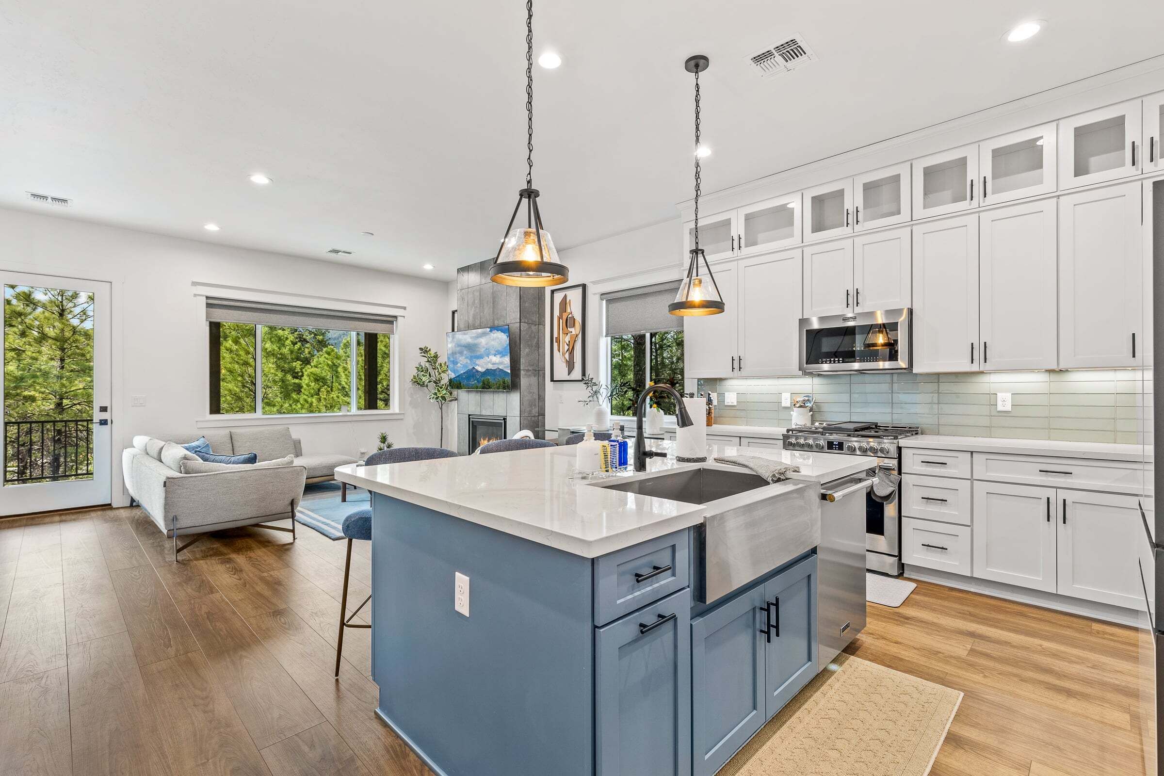 Una cocina moderna con armarios blancos, una isla azul, electrodomésticos de acero inoxidable, iluminación colgante y un amplio ventanal con vistas a árboles.