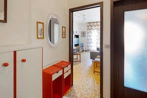 Ein Flur mit einer weißen Tür, einem Spiegel und einem roten Schrank. Im Hintergrund ist ein Zimmer mit einem Bett und einem Fenster erkennbar.