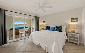 Habitación de hotel con cama, mesita de noche, lámpara, ventilador de techo, vista al mar a través de una puerta corrediza y un cuadro en la pared.
