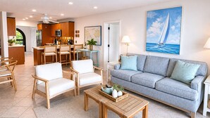 A modern living room with a grey sofa, white armchairs, a wooden coffee table, and a dining area with wooden furniture. - Oceanfront steps to beach & yacht club-generator (St. Thomas)