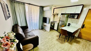 Un espacio habitable compacto con sofá, una mesa de comedor con sillas, zona de cocina con electrodomésticos y ventana con cortinas.