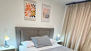 Habitación de hotel con cama, dos mesitas de noche, lámpara, ventana con cortinas y dos obras de arte en la pared.