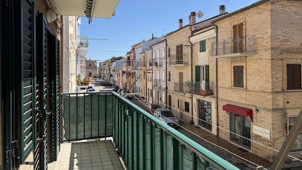 Eine enge Straße, gesäumt von Gebäuden, mit einem Balkon und einem grünen Geländer, und einem wolkenlosen, blauen Himmel.