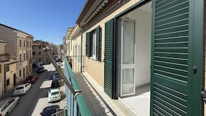 Eine schmale Straße mit parkenden Autos, ein Gebäude mit grünen Fensterläden und einem strahlend blauen Himmel.