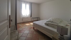 A bedroom with a bed, a bedside table, a lamp, a chair, a window with curtains, and a radiator. - Spacious apartment near Lake Sainte-Croix, surrounded by truffle oaks. (Sainte-Croix-du-Verdon)