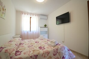 Ein Schlafzimmer mit einem Bett, einem an der Wand montierten Fernseher, einem Fenster mit Vorhängen und einer kleinen Pflanze auf einem Regal.