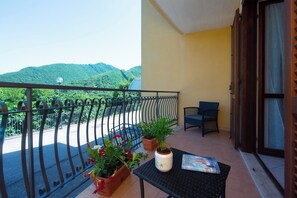 A balcony with a view of green hills, a potted plant, a chair, and a table with a book. - Sunsets apartment (Pietre)