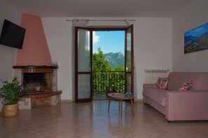 A living room with a fireplace, a sofa, a table, and a balcony with a mountain view. - Sunsets apartment (Pietre)