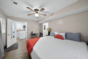 A hotel room with a bed, a desk, a chair, a ceiling fan, and a laundry area.