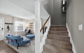 A staircase leading to an upper floor, a living room with a blue sofa and ottoman, and a wall-mounted shelf with decorative items.