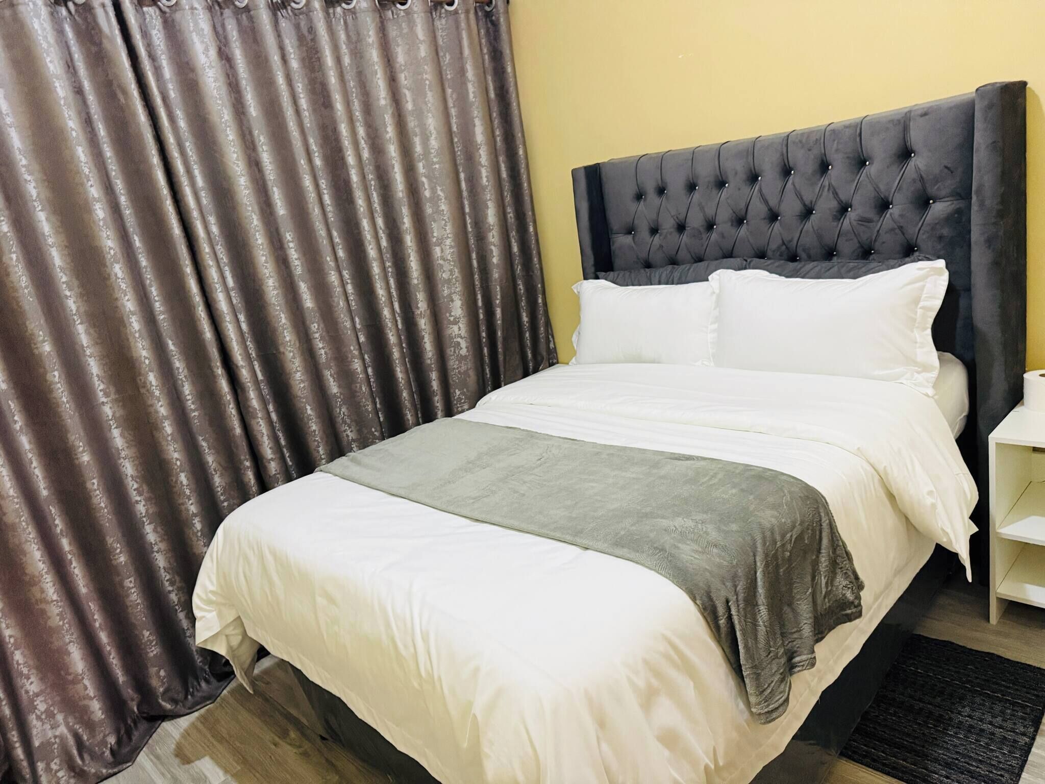 Una cama bien hecha con ropa de cama blanca y gris, un cabecero capitonado y una cortina oscura.