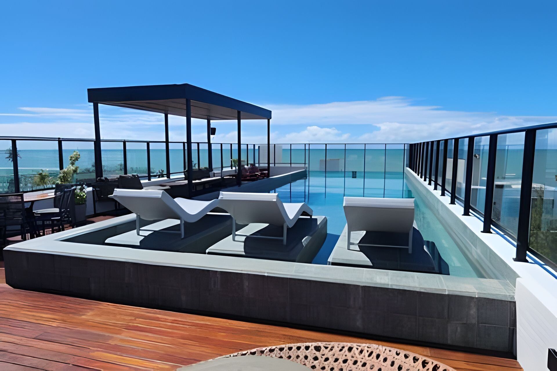 A rooftop pool area with lounge chairs, a covered seating area, and a glass balustrade overlooking the sea.