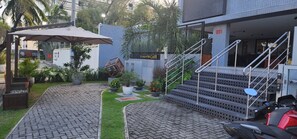 A paved pathway leading to a building entrance with a staircase, surrounded by potted plants and a palm tree. - Flat Beira mar João Pessoa (João Pessoa)