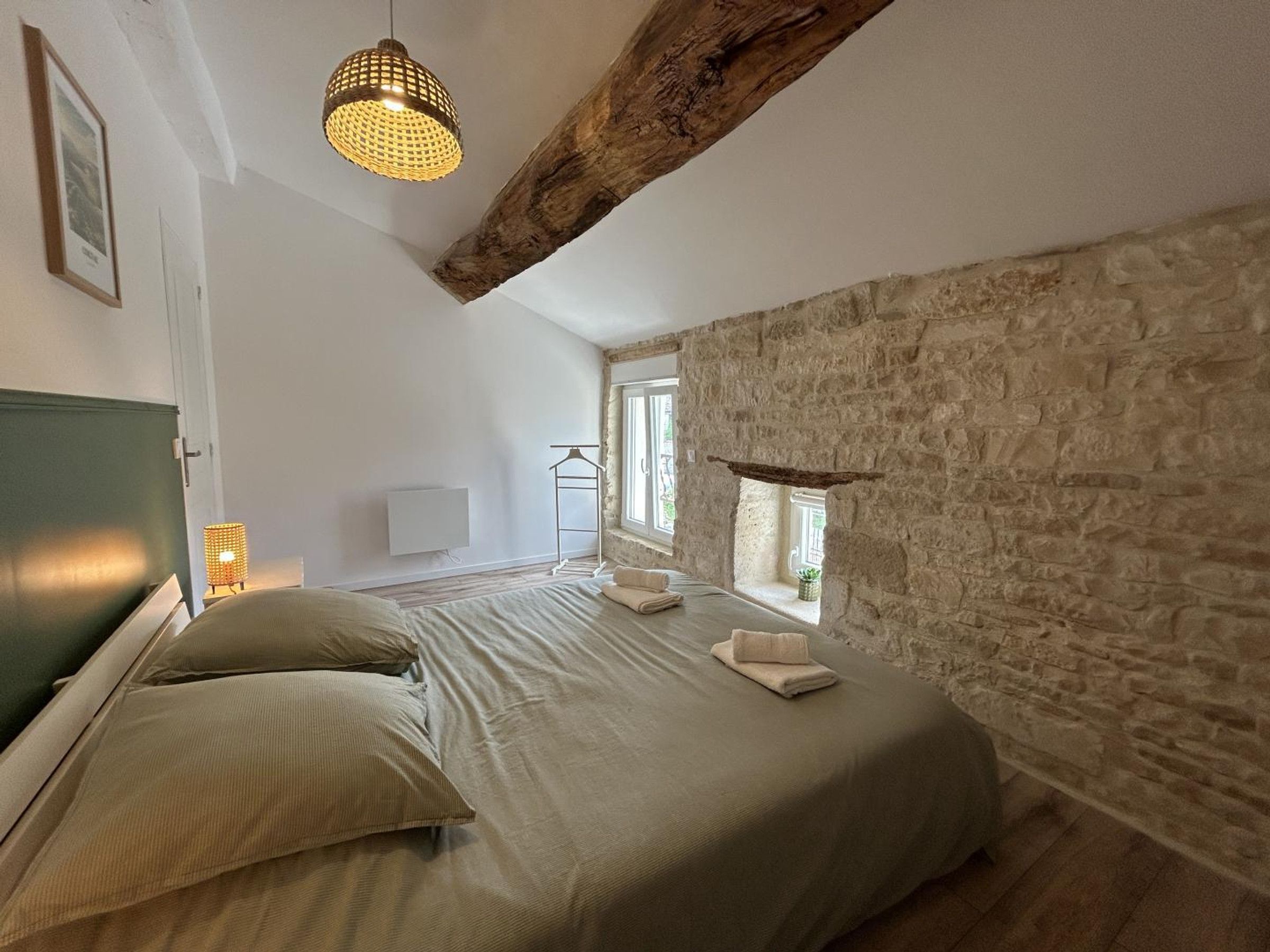 A bedroom with a large bed, a stone wall, a wooden beam ceiling, and a hanging light fixture.