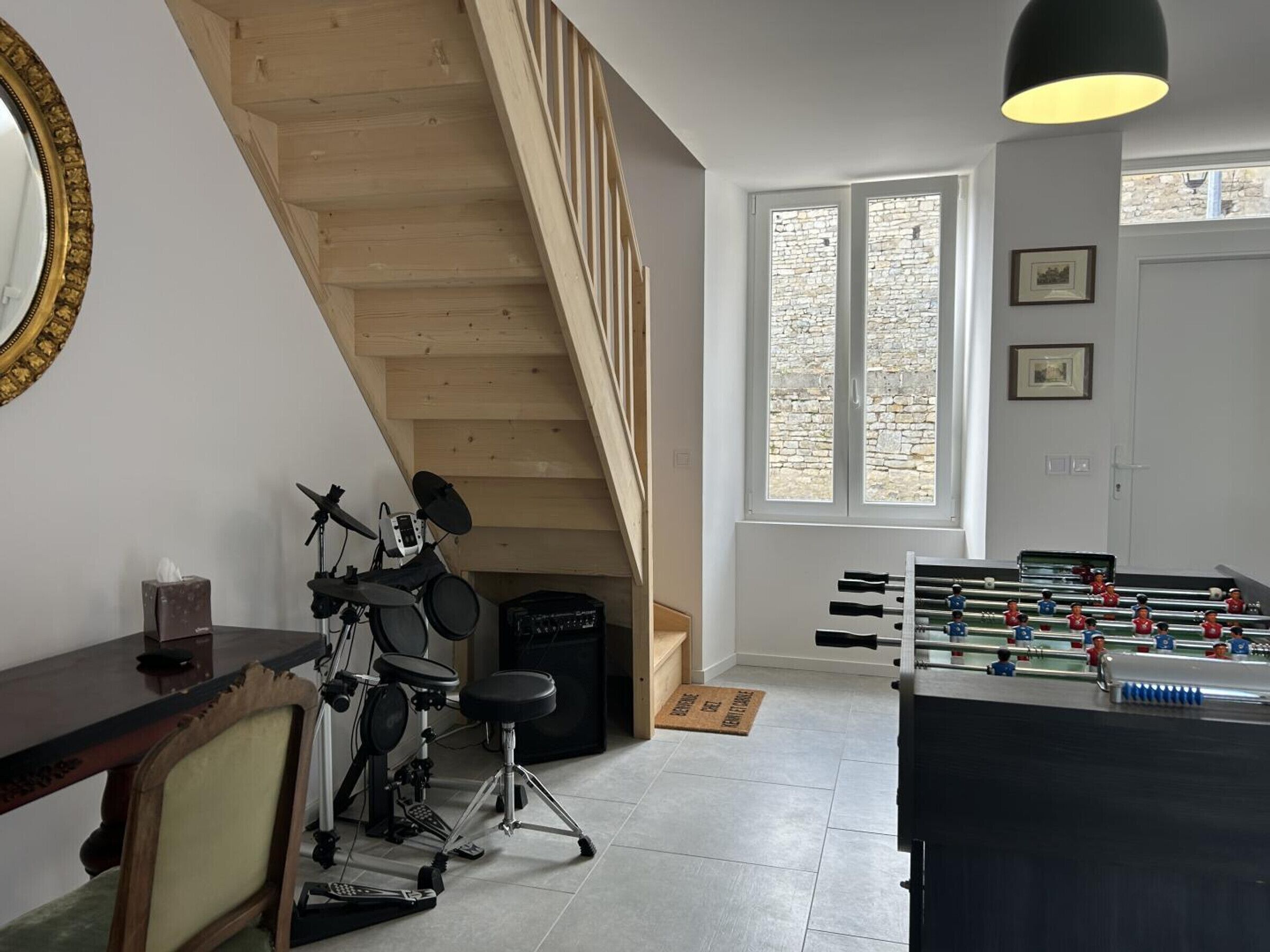 Une pièce équipée d’une batterie, d’un baby-foot, d’un escalier et d’une fenêtre donnant sur un mur en pierre.
