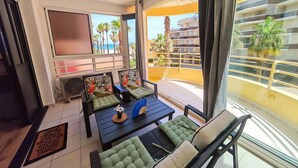 Un balcón con vista a palmeras y un edificio, un sofá con cojines verdes, una mesita con un ventilador y una puerta con felpudo.