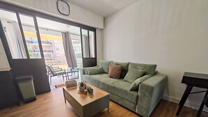 Una sala de estar con un sofá verde, una mesa de centro de madera, un cubo de basura y vistas a través de ventanas grandes.