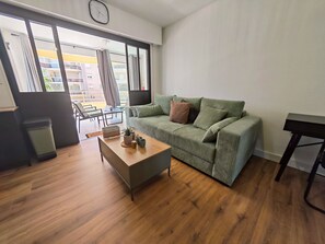 Ein Wohnzimmer mit einem grünen Sofa, einem Holztisch, einem Mülleimer und Blick durch große Fenster.