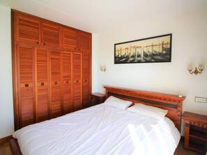 A bedroom with a large wooden wardrobe, a bed with white linens, a nightstand, a wall-mounted lamp, and a framed picture of boats. - Apartment with AC, sea view - 4 guests (Roses)