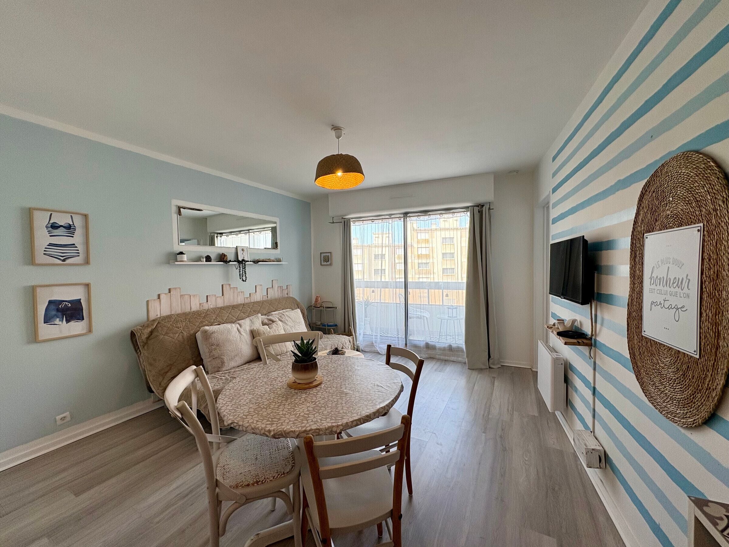Una sala de estar con un sofá, una mesa de comedor con sillas, un televisor montado en la pared y un ventanal con vistas a edificios.