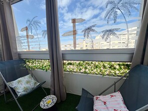 A balcony with two chairs, a small table, and a view of palm trees and buildings. - Charming apartment - 4 guests, sea view (Saint-Hilaire-de-Riez)