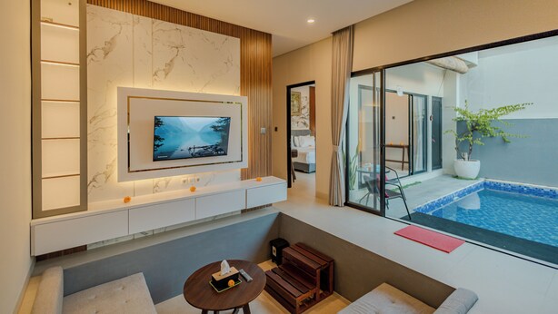 A modern hotel room with a TV mounted on a marble wall, a small seating area, and a view of a swimming pool. - Shrivatsa Home Villa (Munggu)