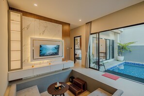 A modern hotel room with a TV mounted on a marble wall, a small seating area, and a view of a swimming pool. - Shrivatsa Home Villa (Munggu)