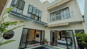 A modern multi-story building with a balcony, large windows, and a visible bedroom with a bed and curtains. - Shrivatsa Home Villa (Munggu)