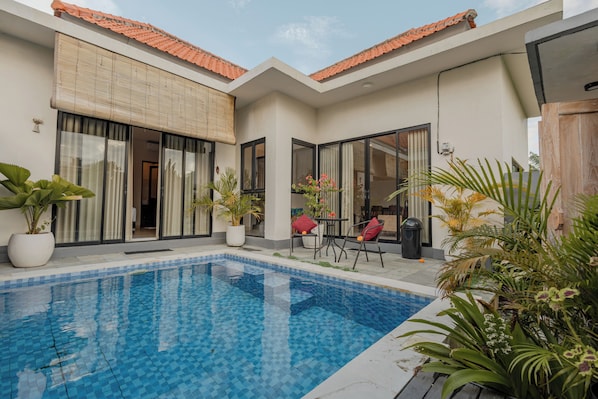 A swimming pool with tiled flooring, a patio area with chairs and potted plants, and a building with a tiled roof and large windows.
