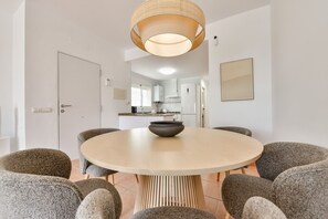 Ein moderner Essbereich mit einem runden Tisch, sechs grauen Stühlen, einer Schale in der Mitte sowie einer Hängeleuchte. Im Hintergrund ist eine Küche zu sehen.