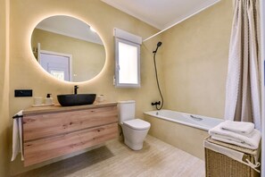 Une salle de bain avec un meuble-lavabo en bois, un miroir rond, une baignoire et des toilettes.