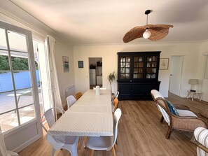 A dining area with a table and chairs, a black cabinet, a sofa, and a hanging light fixture. - Calm villa in Cap-Ferret - 7 guests (Lège-Cap-Ferret)