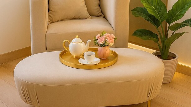 Un salon moderne avec un canapé, un pouf rond, un plateau contenant une théière, une tasse et des fleurs, ainsi qu’une plante en pot.