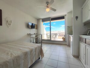 Ein Hotelzimmer mit Bett, Fernseher, Deckenventilator, Balkon mit Stühlen und Blick auf den Himmel.