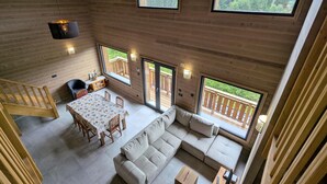 Una cabaña de madera con techos altos, comedor, sofá y ventanas grandes que ofrecen vistas de una montaña boscosa.
