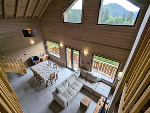 Une cabane en bois aux plafonds hauts, comprenant un coin repas, un canapé et de grandes fenêtres donnant sur une montagne boisée.