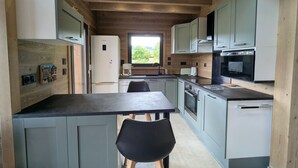 Una cocina moderna con vigas de madera, una zona de comedor con una mesa y sillas, y vistas a zonas verdes al exterior.