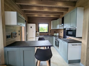 Une cuisine moderne avec des poutres en bois, un coin repas avec une table et des chaises, et une vue sur la verdure à l’extérieur.