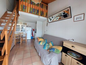 A compact living space with a sofa, wooden staircase, kitchen area, and a mirror reflecting the room. - Apartment in Vaux-sur-Mer - 4 guests (Vaux-sur-Mer)