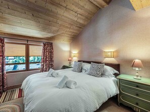 A bedroom with a wooden ceiling, a large bed with white linens, two bedside lamps, and a view through a window with patterned curtains. - Apartment in the center - 10 guests (Méribel)