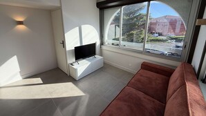 Una sala de estar moderna con un televisor sobre un mueble blanco, un ventanal con vistas a edificios y un sofá rojo.