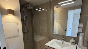 Un baño moderno con ducha de vidrio, un lavamanos blanco sobre un mueble de madera y un espejo grande.