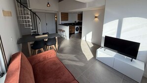 A modern living room with a sofa, a flat-screen TV, and a staircase leading to a kitchen area. - Duplex apartment, air conditioning, central - 4 guests (Port Leucate)