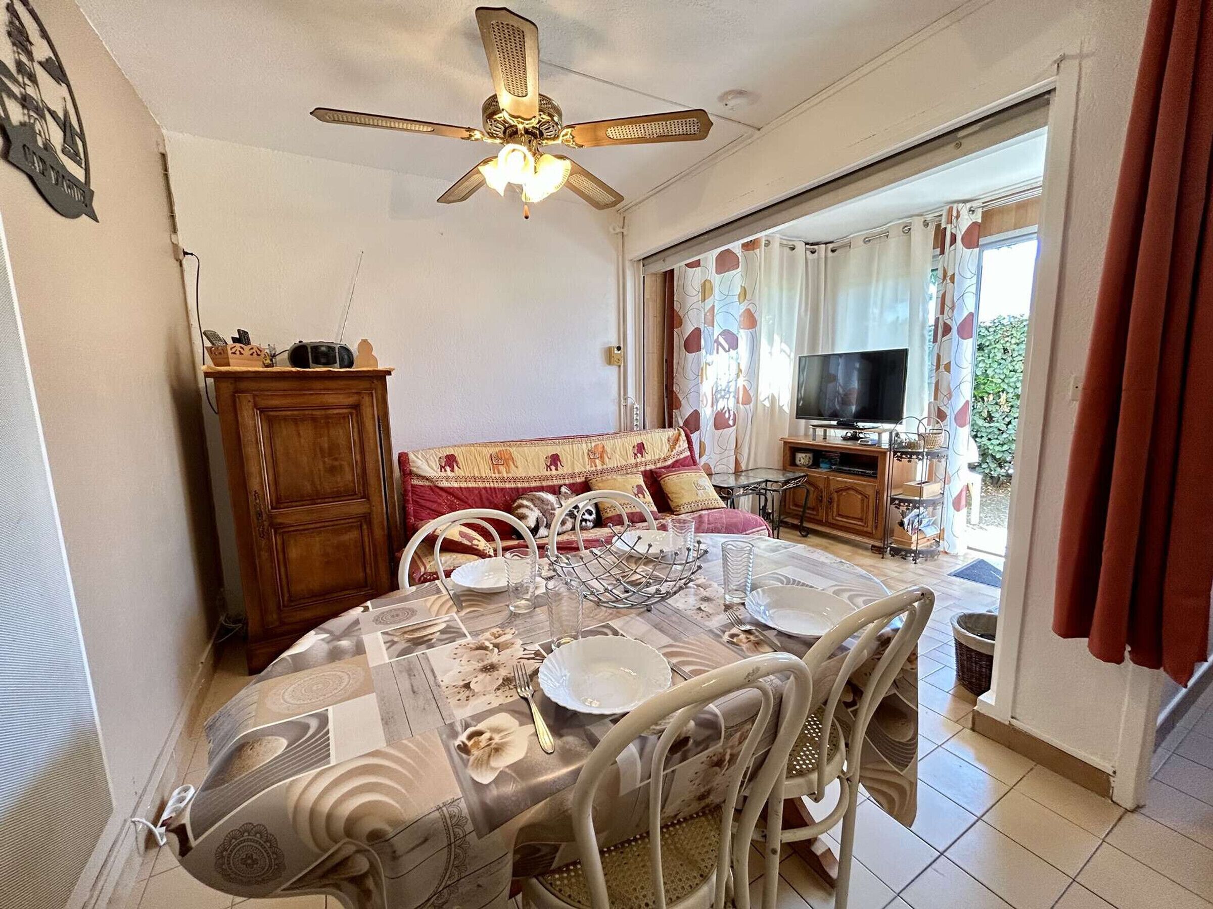 A dining area with a table set for four, a ceiling fan, a sofa with patterned cushions, and a television in the background.