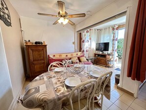Ein Essbereich mit einem Tisch, gedeckt für vier Personen, einem Deckenventilator, einer Couch mit gemusterten Kissen und einem Fernseher im Hintergrund.