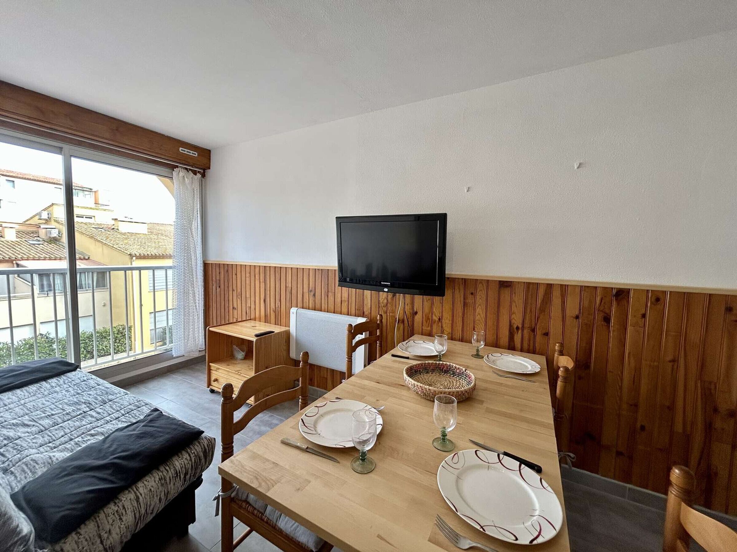 A room with a wooden dining table set for four, a television mounted on the wall, and a bed with a black headboard.