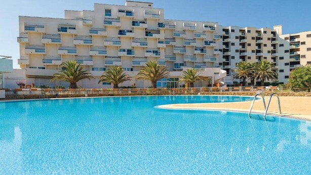 A swimming pool with a clear blue water surface, surrounded by a building with multiple balconies and palm trees.